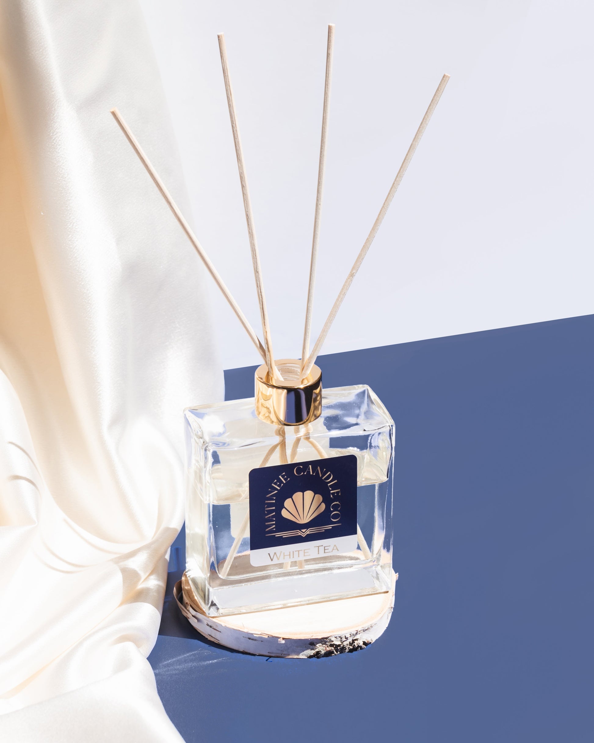 The classic reed diffuser in White Tea on display next to an ivory satin sheet against a blue and white background